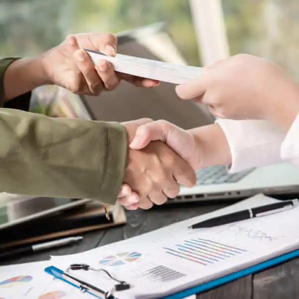 Close-up of two people shaking hands while exchanging a stack of documents or cash over a desk with financial charts.